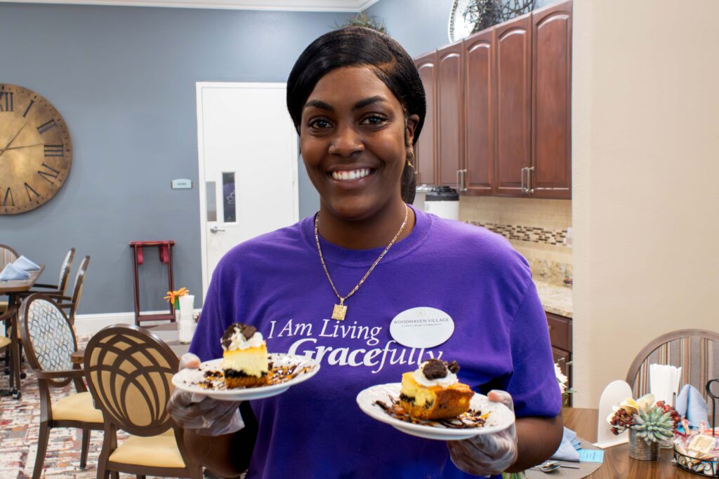 A staff member stands in the kitchen holding two plates of dessert.