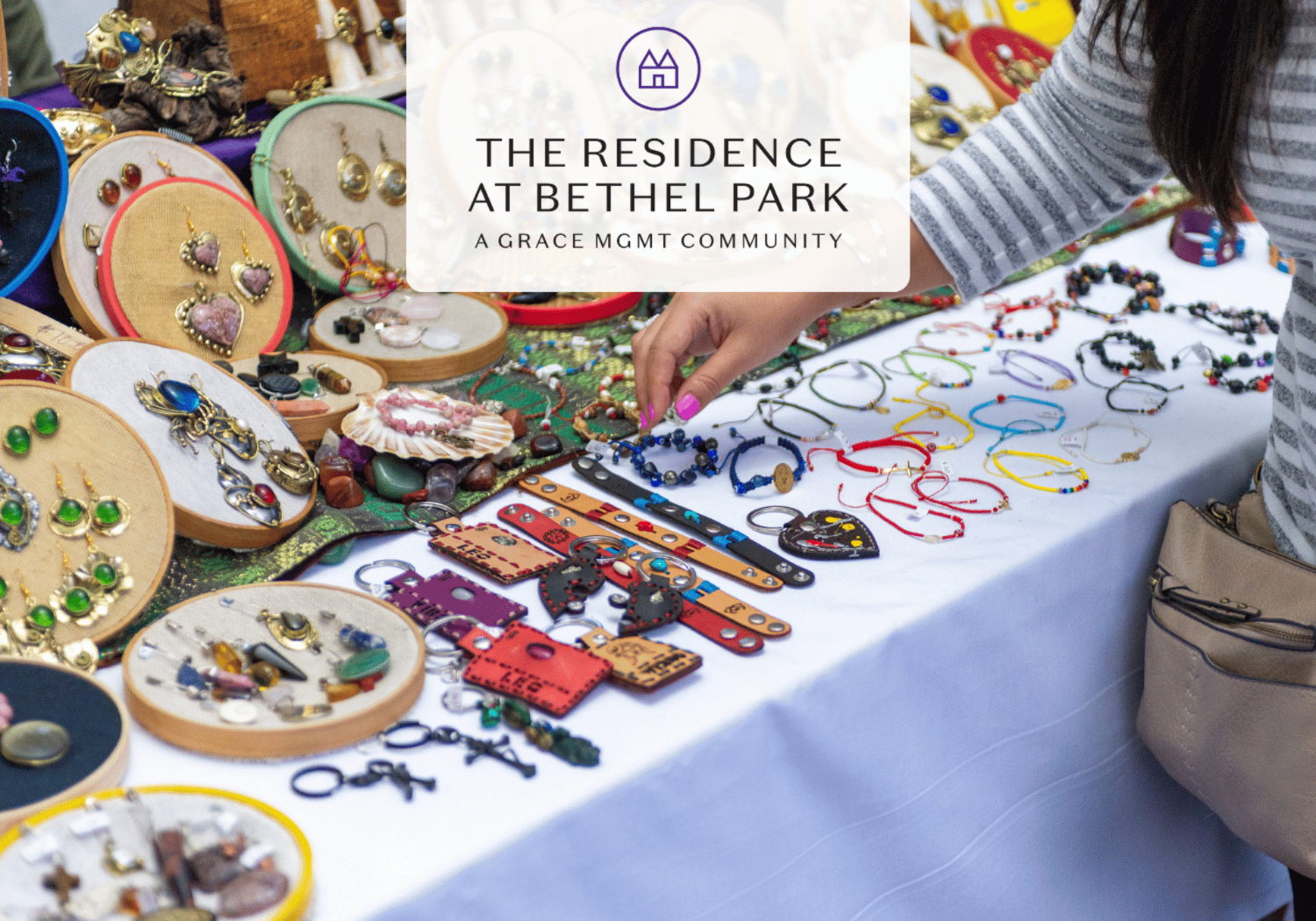 Market table filled with colorful jewelry including bracelets and earrings; a person’s hand selects a blue beaded bracelet.