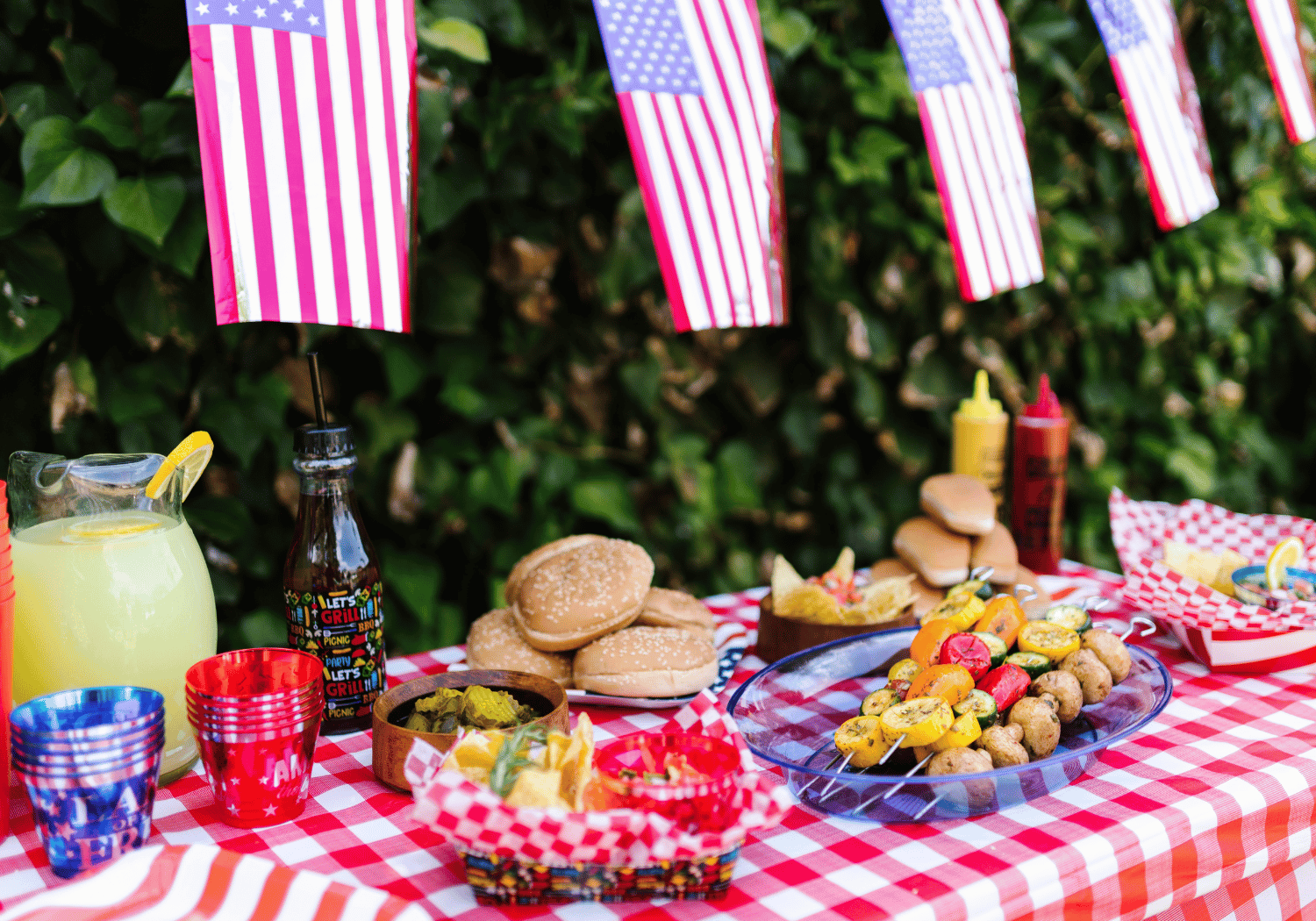 Outdoor 4th of July picnic table with red‑and‑white checkered cloth, American flags, lemonade, burgers, chips, and colorful skewered veggies.